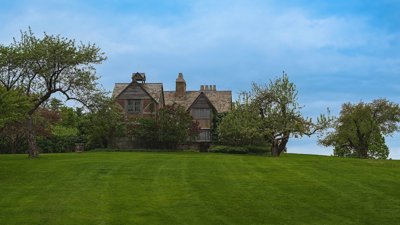 A stone house with a gabled roof sits behind green trees on a large, well-manicured lawn under a blue, partly cloudy sky.