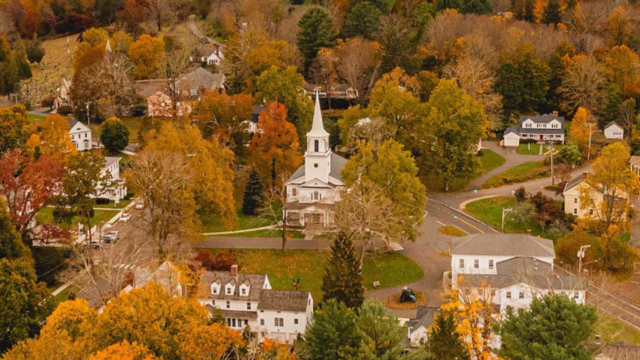 Aerial view of a small town with a white church, autumn trees with colorful leaves, and scattered houses.