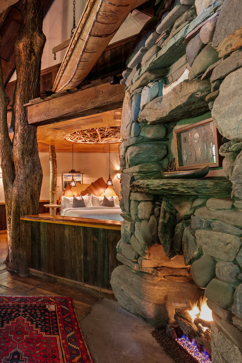 Rustic bedroom with stone fireplace, exposed wood beams, hanging lights, and a bed with white pillows and wooden headboard; vibrant rug on floor.