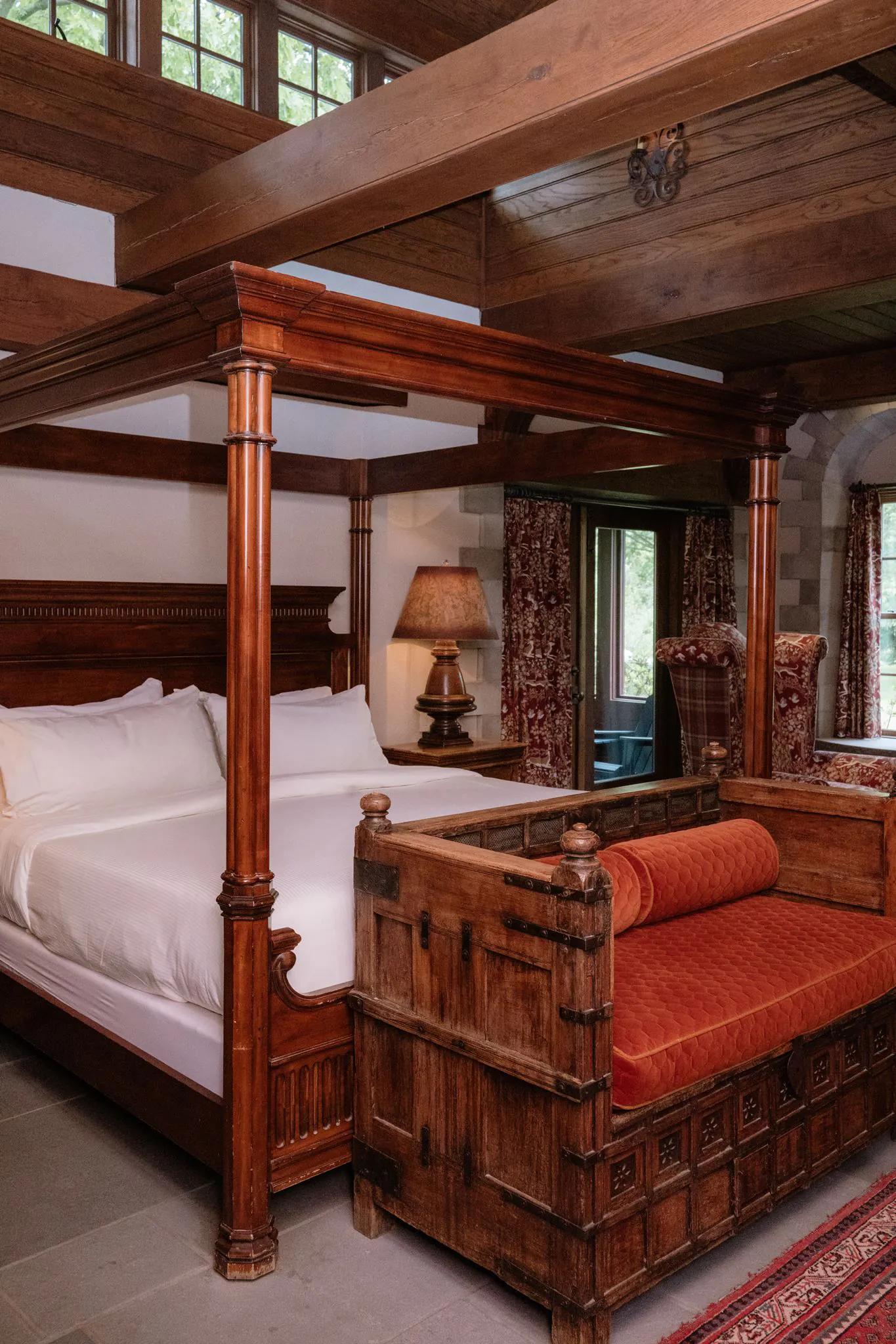 A cozy bedroom with a wooden four-poster bed, white bedding, a wooden chest, red cushions, a lamp, and large windows with patterned curtains.