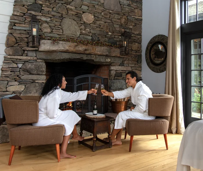 Couple in white bathrobes toasting with champagne glasses by a stone fireplace