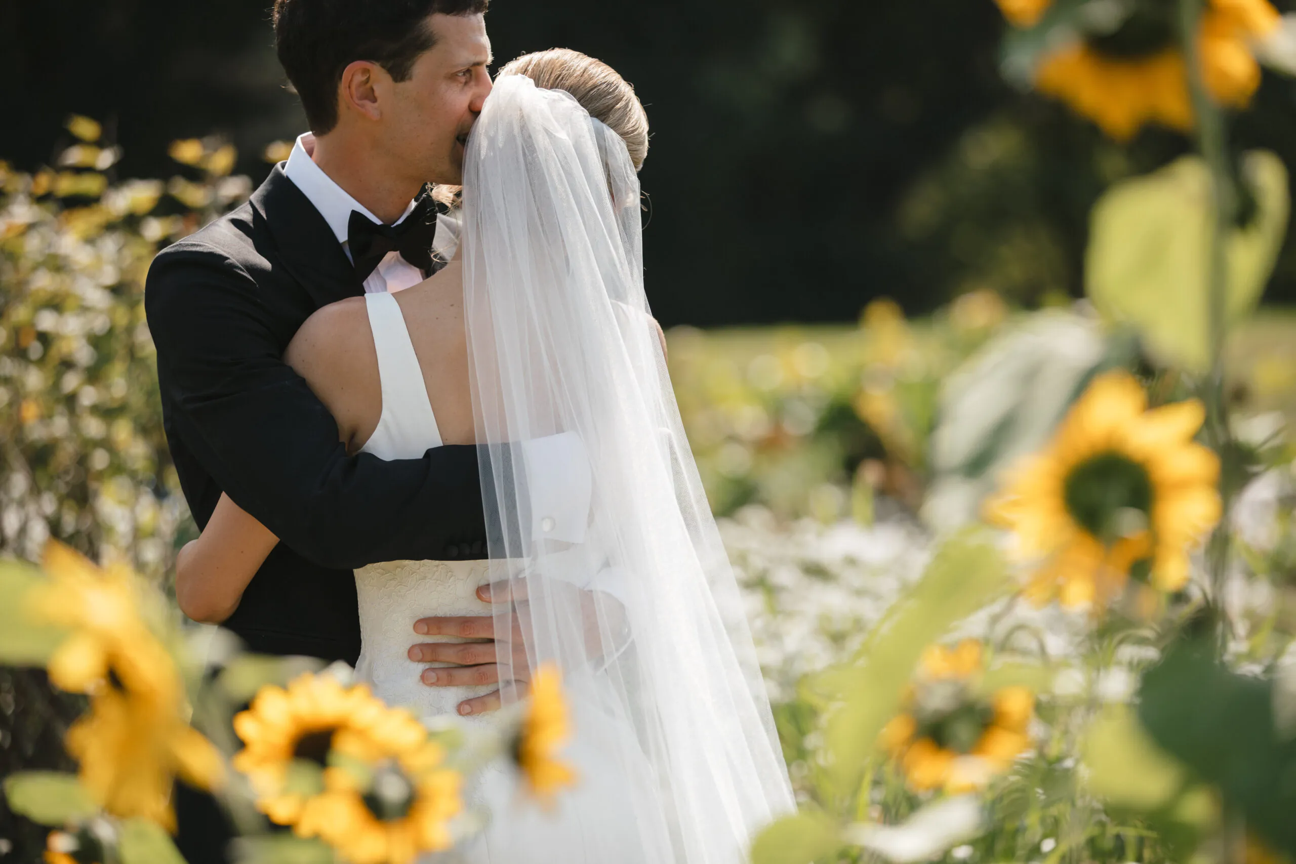 A groom in a black suit kisses his brides forehead as they embrace in a sunny garden surrounded by blooming sunflowers.