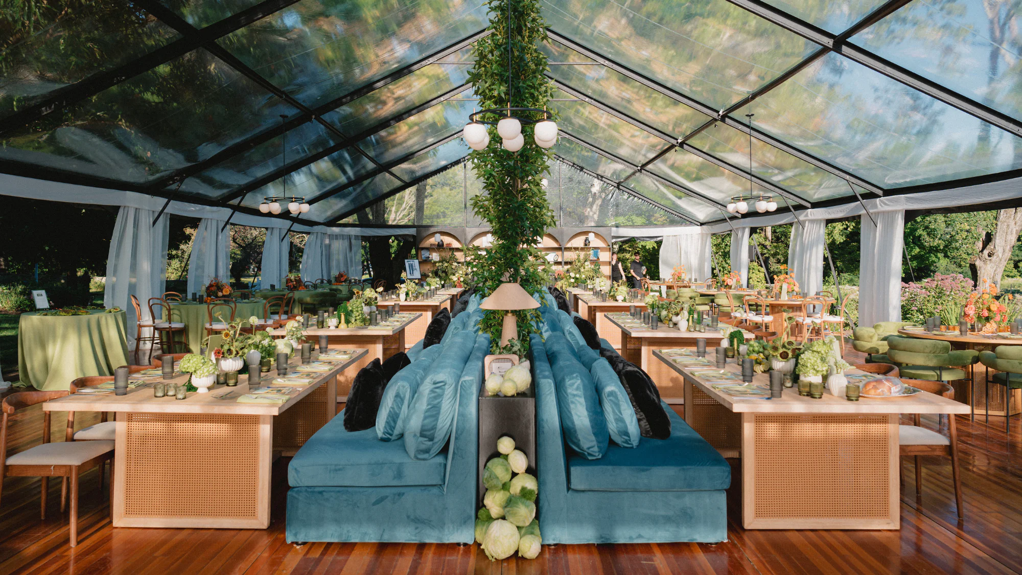 A clear-roofed event tent set up for dining with long wooden tables, blue velvet booths, and green floral centerpieces.