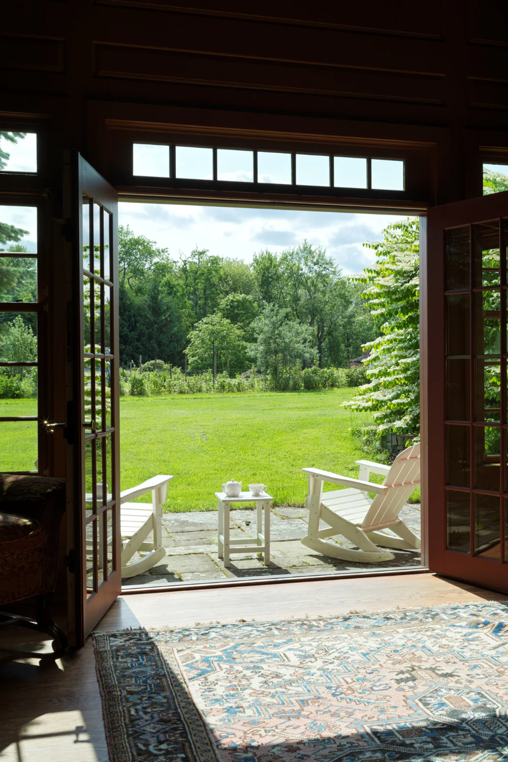 Open double doors reveal a green lawn with two white Adirondack chairs and a small table on a stone patio, seen from inside a cozy room.