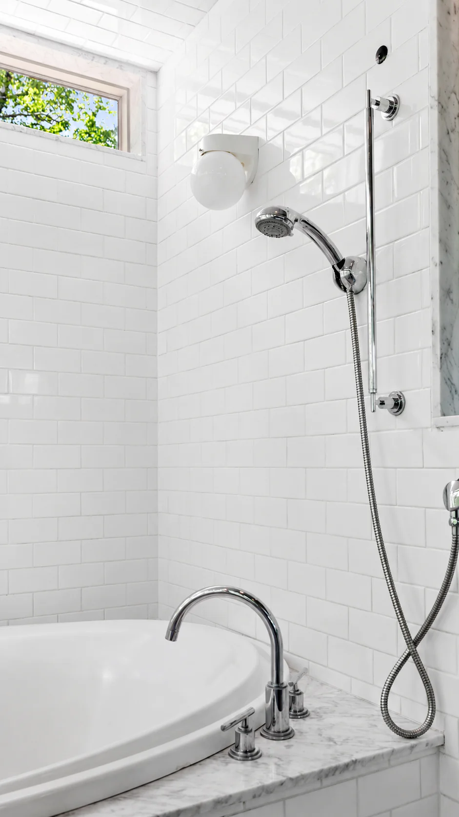 White tiled bathroom with a bathtub, chrome faucet, handheld showerhead, wall light, and a small window showing green leaves outside.