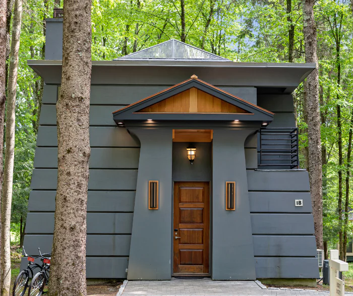 Modern gray house with a triangular roof detail, wooden door, and lantern, surrounded by tall trees; two bikes are parked beside the entrance.