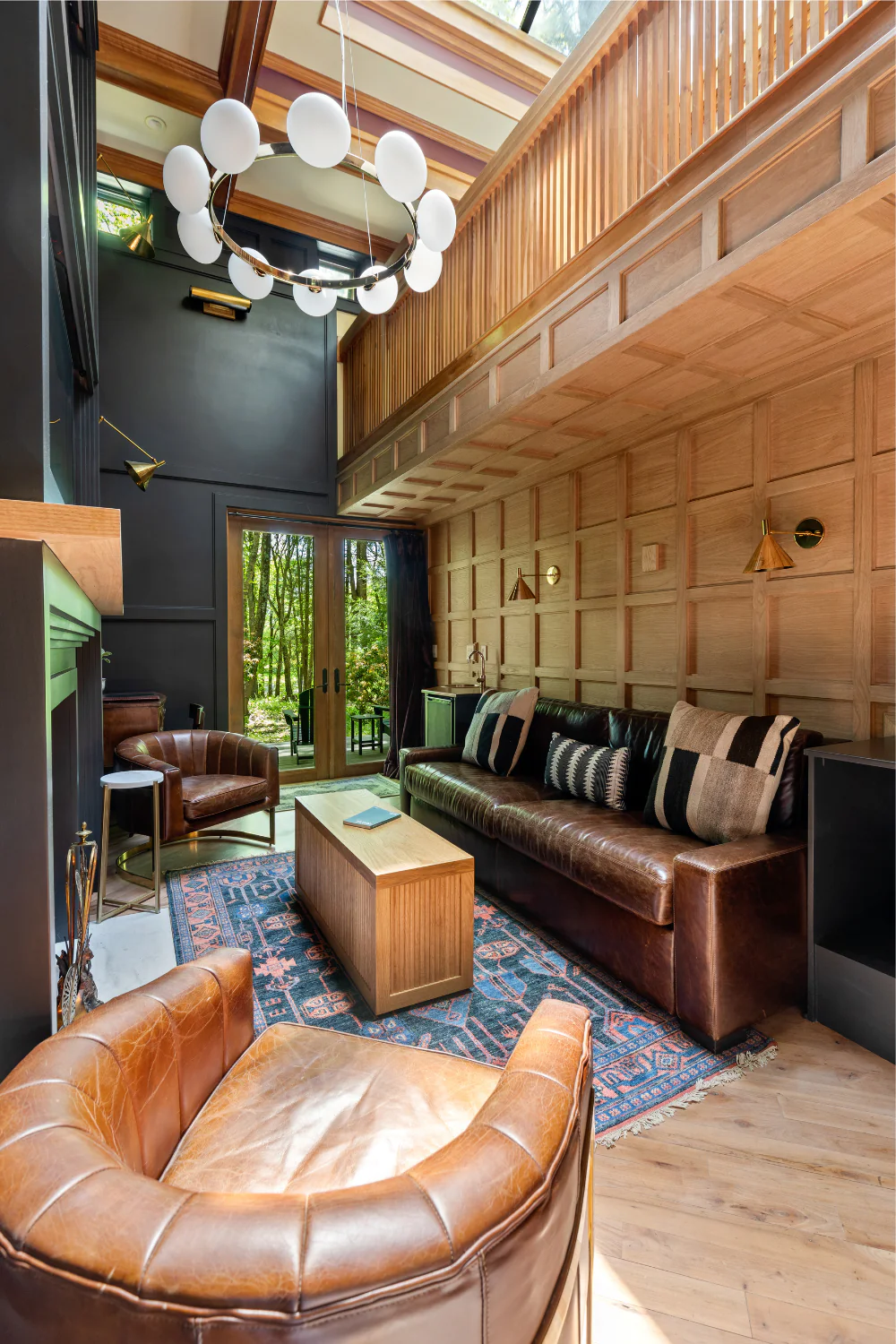 Modern living room with leather chairs, black sofa, wood paneling, a rug, large chandelier, and floor-to-ceiling windows showing trees outside.