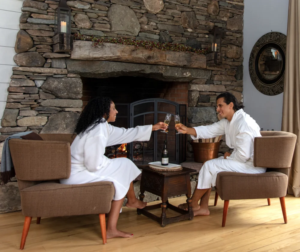 A couple in robes toasting with champagne by a stone fireplace.