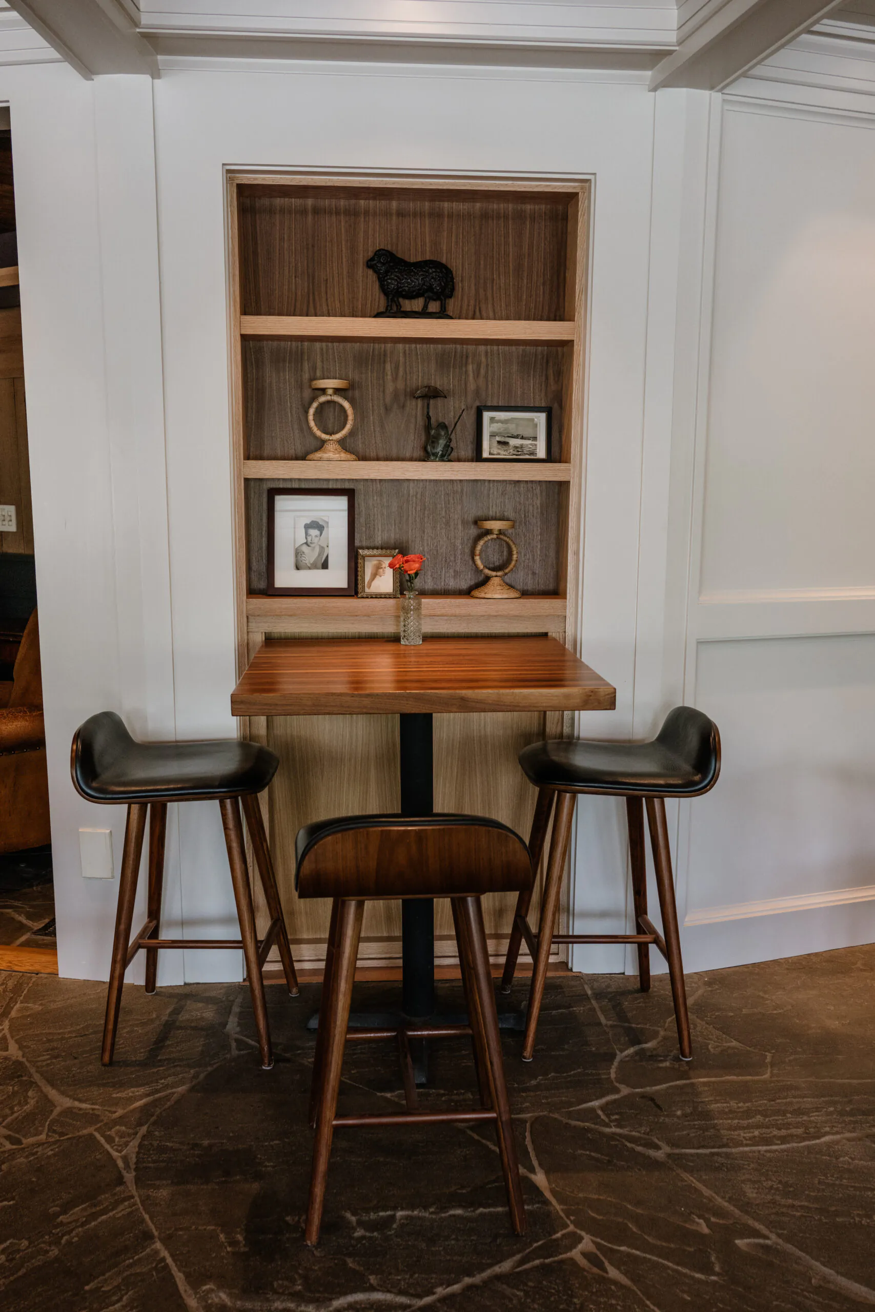 A small wooden table with two barstools, a single flower in a vase, and built-in shelves displaying framed photos and decorative objects.