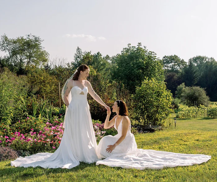 Two brides in white wedding dresses in a garden, one kneeling and holding the others hand.