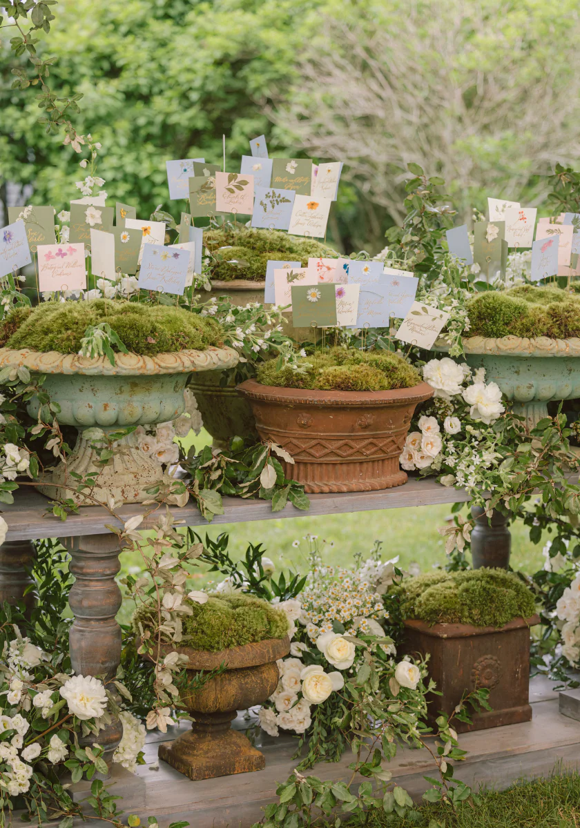 Escort cards with handwritten names displayed on moss-filled urns arranged on a wooden table decorated with white flowers.