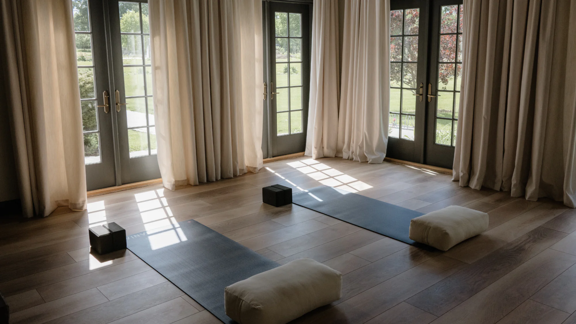 Two yoga mats on a wood floor, each with a foam block and a bolster, in front of glass doors with curtains.