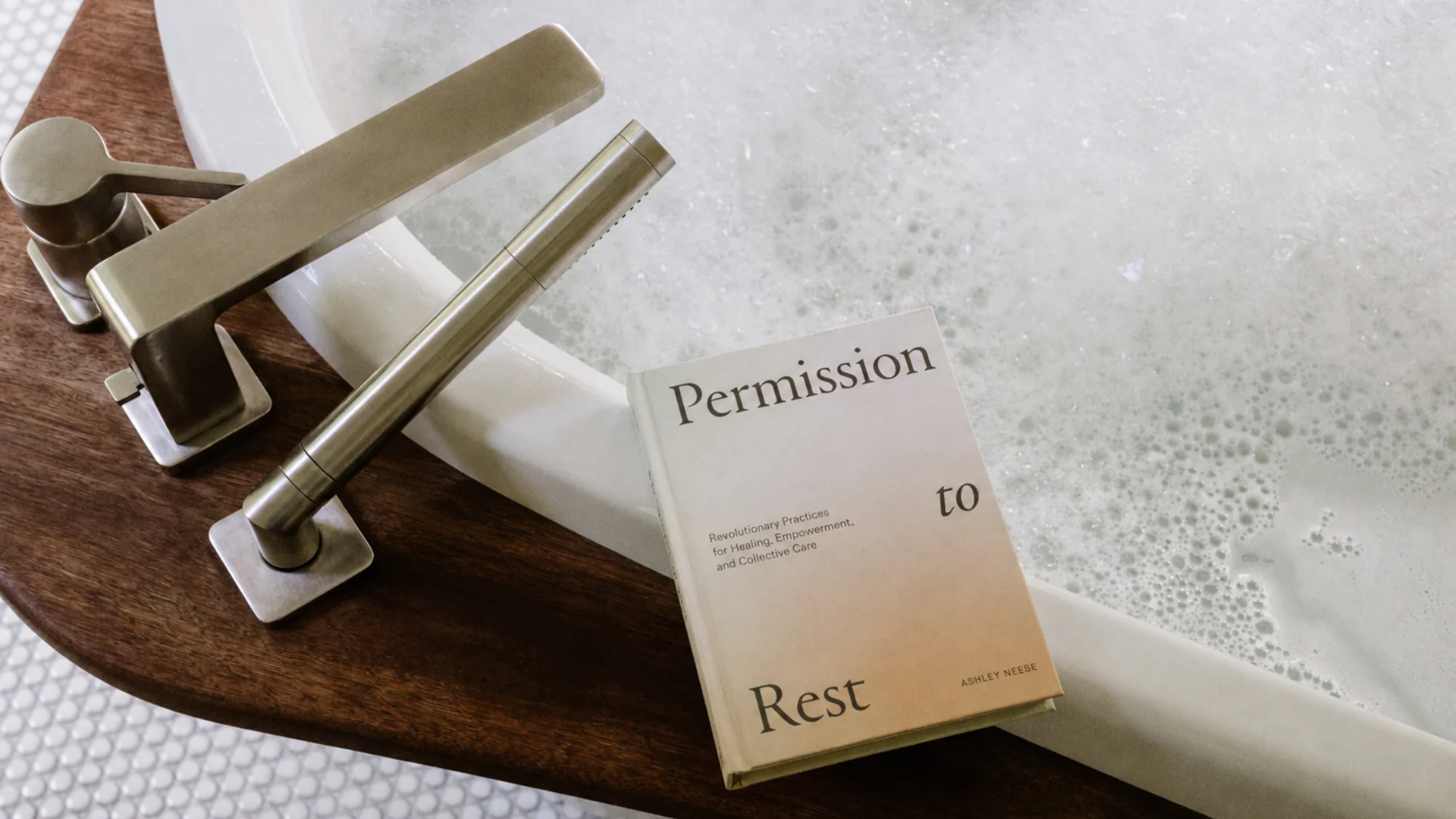 The book Permission to Rest by Ashley Neese resting on the edge of a bubble-filled bathtub.