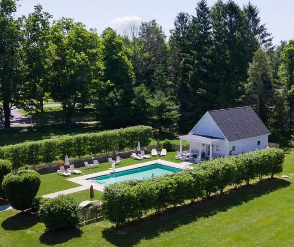 Aerial view of a rectangular swimming pool surrounded by lounge chairs and a white pool house, set within a lush green yard with tall trees.