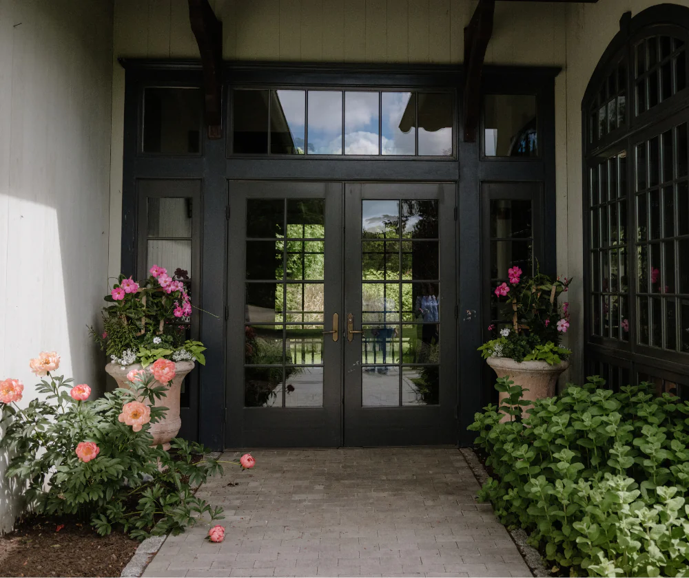 Double glass doors with a black frame, flanked by stone planters with blooming pink peonies and lush green foliage on a brick walkway.