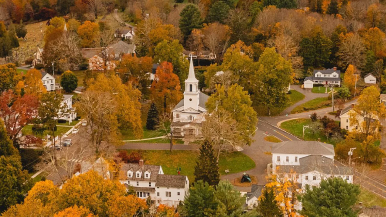 Aerial view of a small town with a white church, autumn trees with colorful leaves, and scattered houses.