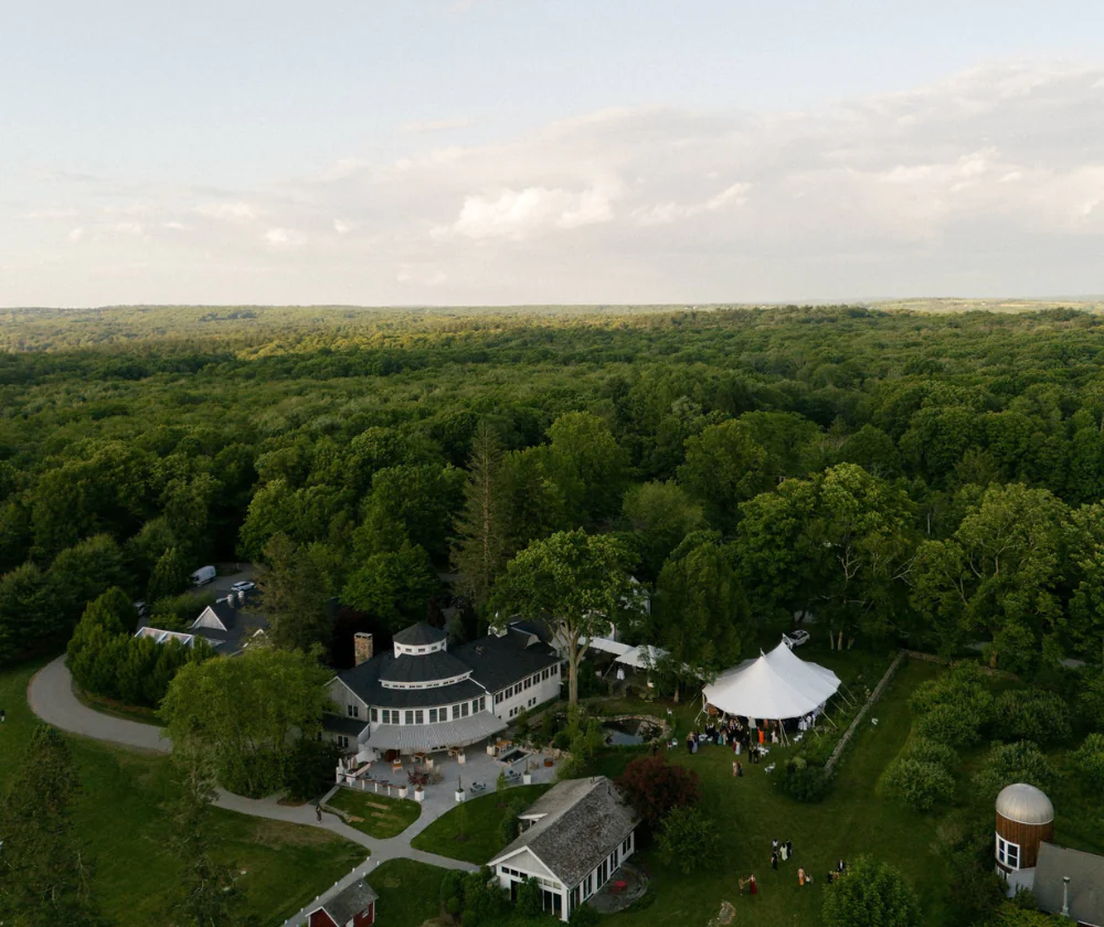 Aerial view of a large estate with a circular building and a white event tent nestled among dense forest.