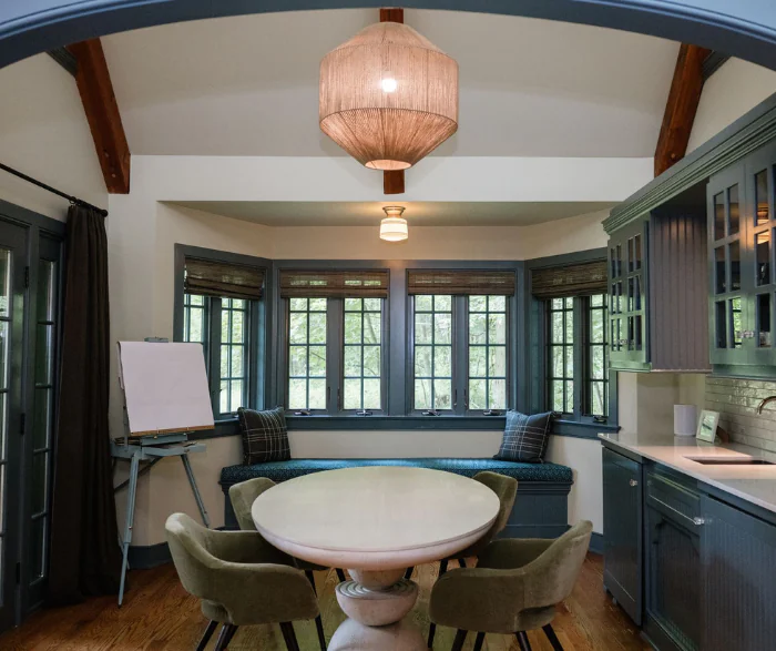 A cozy dining nook with a round table, four green chairs, window bench seating, large windows, and a pendant light above.