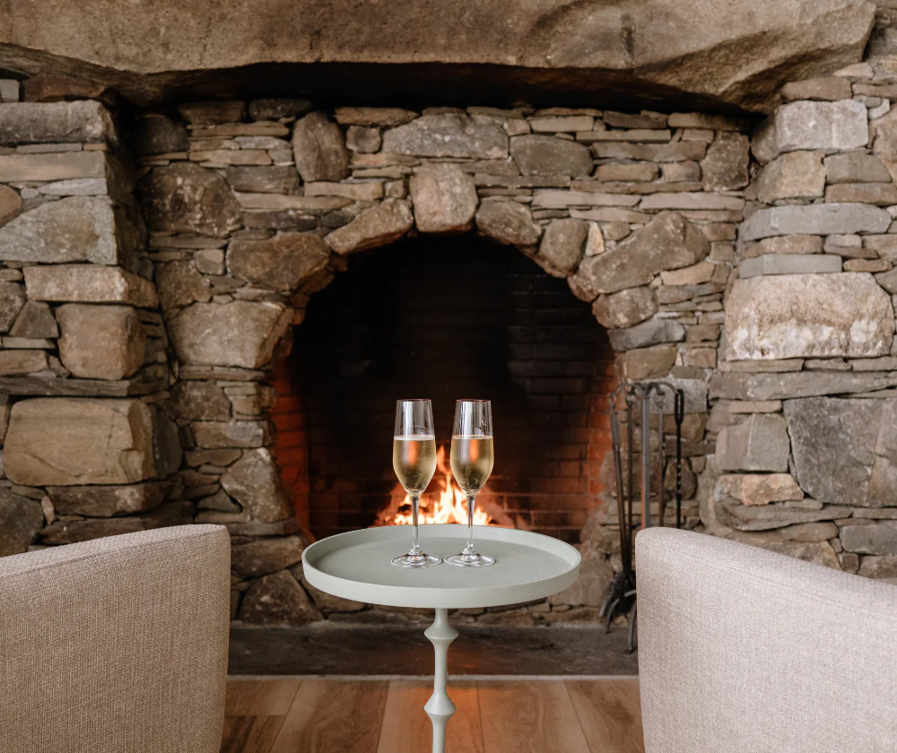 Two champagne glasses on a small table in front of a stone fireplace with a fire burning, flanked by two armchairs.
