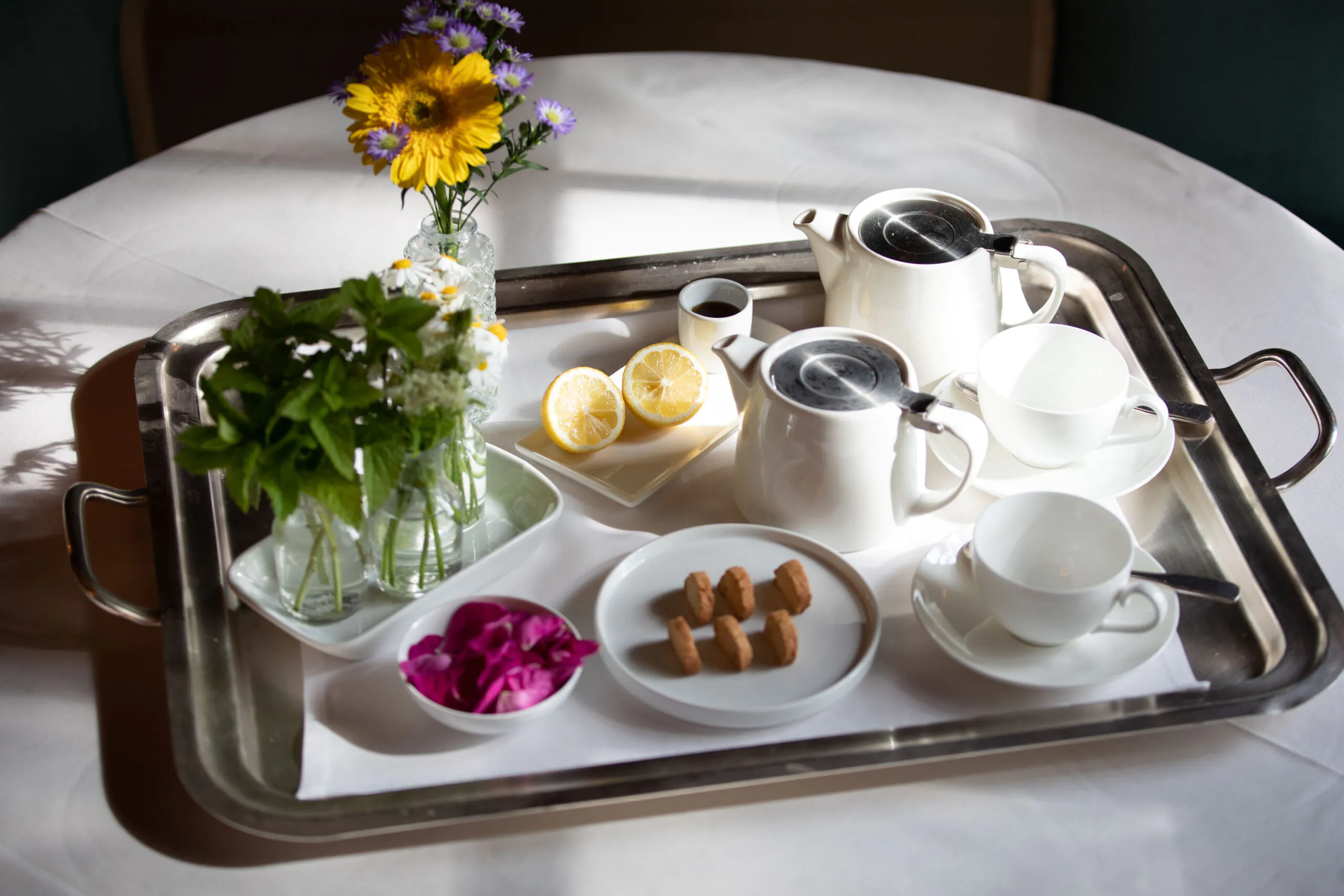 A silver tray with a teapot, teacups, lemon slices, sugar cubes, flowers in vases, and a small pitcher, set on a white tablecloth.
