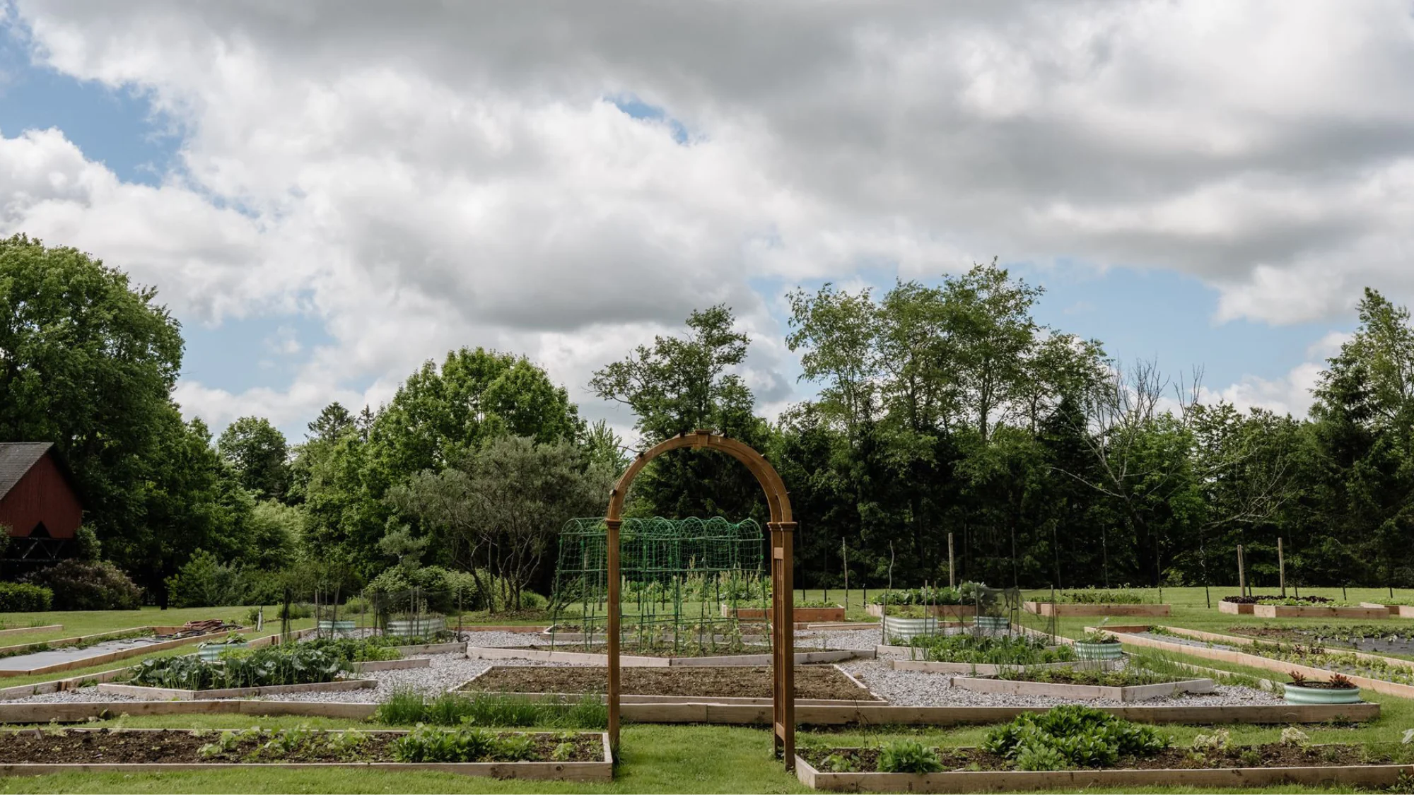 A large garden with raised beds, a wooden arch, and green plants, surrounded by grass and trees under a cloudy sky.