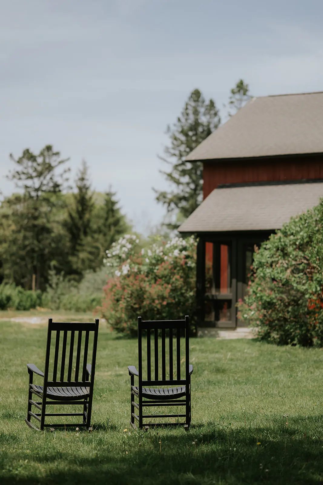Two black rocking chairs sit on a grassy lawn near a red wooden house, surrounded by green trees and bushes under a blue sky.