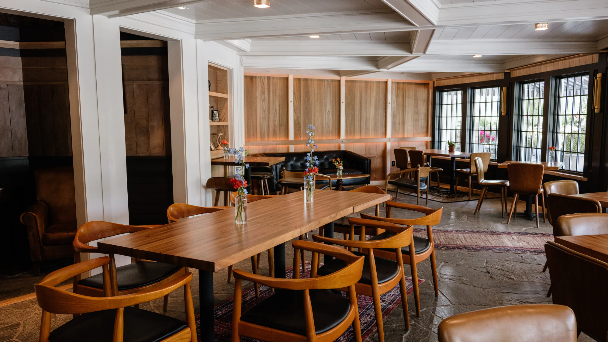 Modern dining area with wooden tables and chairs, vases with flowers, large windows, and built-in wooden cabinets along the walls.