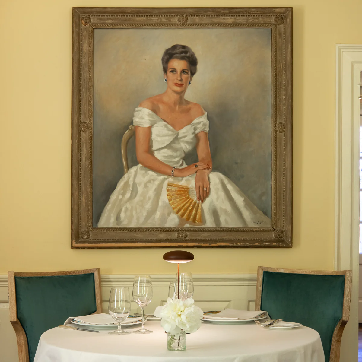 A formal dining table with green chairs and a flower centerpiece sits beneath a large framed portrait of a woman in a white gown holding a fan.