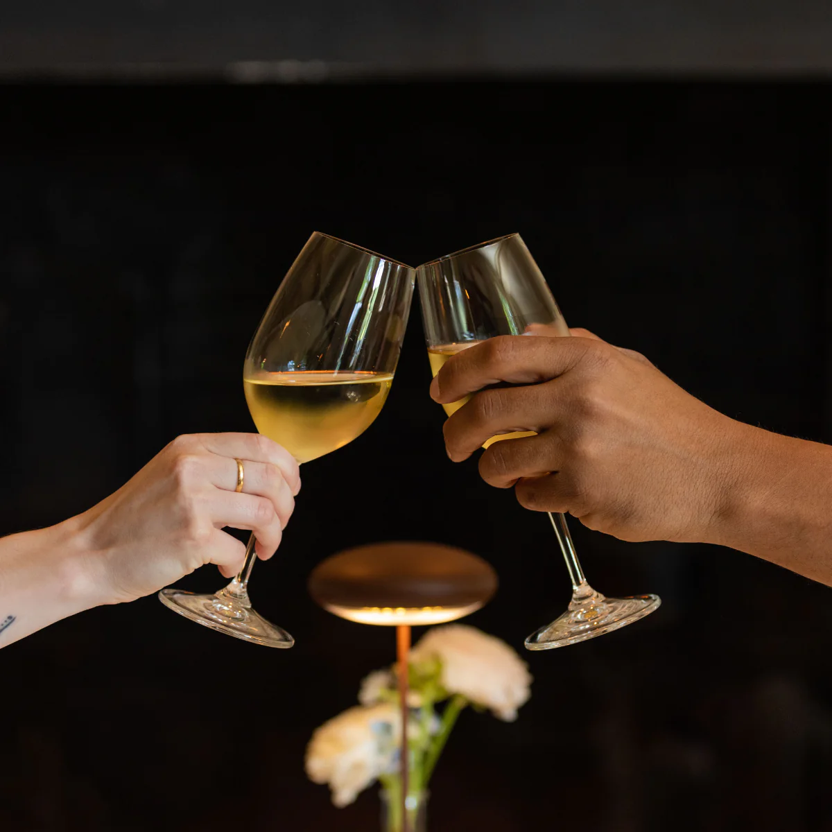 Two hands clink glasses of white wine over a blurred background with flowers and a lamp.