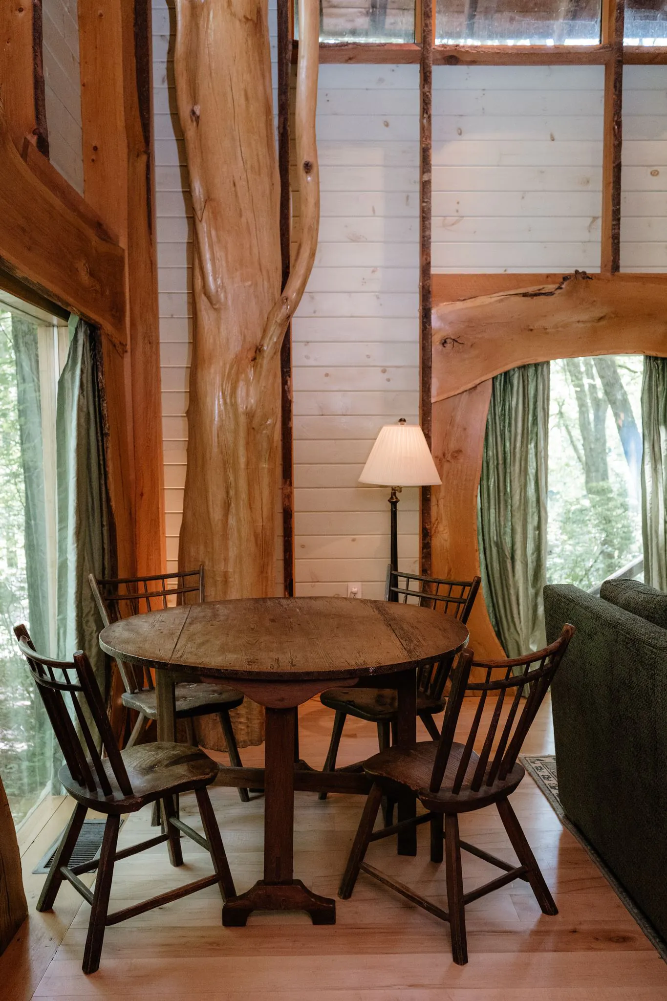 A rustic wooden dining table with four chairs sits by large windows in a cozy cabin with tree trunk beams and natural wood accents.